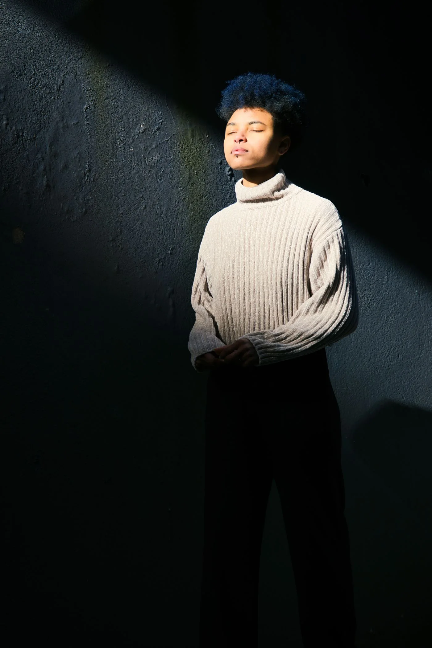 A person in a gray turtleneck sweater standing in sunlight near a wall.